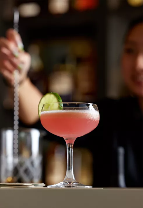 A pink cocktail with a lime wedge on the side of the glass set atop the Mary Mary Bar with a unfocused bartender behind