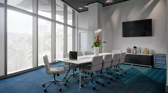 a conference room with floor to ceiling windows and a white conference table in the middle, surrounded by while rolling chairs