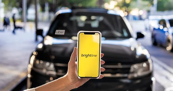 A smart phone with the Brightline app being held up in front of a rideshare vehicle with an uber placard in the windshield.