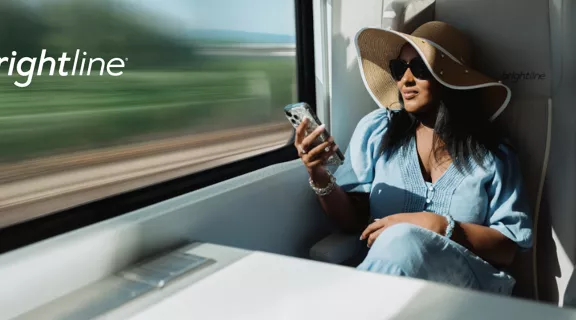 A woman sits by the window on a modern train, looking at her smartphone while traveling. She wears a wide-brimmed hat and casual clothing, suggesting a relaxed journey. The blurred green landscape outside emphasizes motion and speed. The text "amadeus x brightline" appears prominently on the left side of the image.