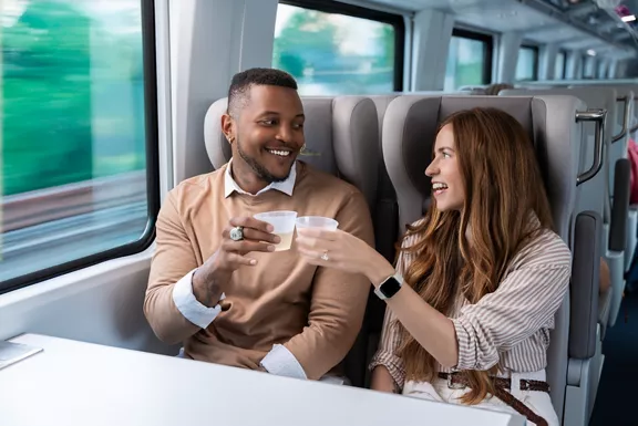 A man and woman toasting their drinks while taking a ride on a Brightline train.