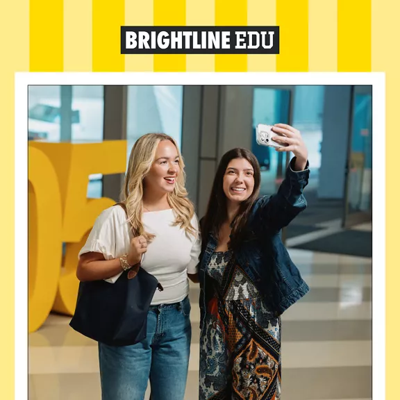 Two young women stand together indoors, smiling as one holds up a smartphone to take a selfie in a Brightline train lobby.