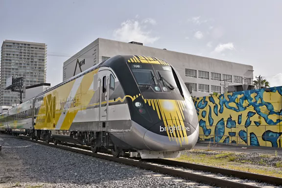 A Brightline passenger train travels along railroad tracks in an urban area. The train features a sleek silver body with bold yellow graphic accents and the word "brightline" visible on the front. Colorful street art covers a wall beside the tracks, while modern high-rise and mid-rise buildings rise in the background. The scene is captured in daylight under a partly cloudy sky, emphasizing contemporary city transportation.