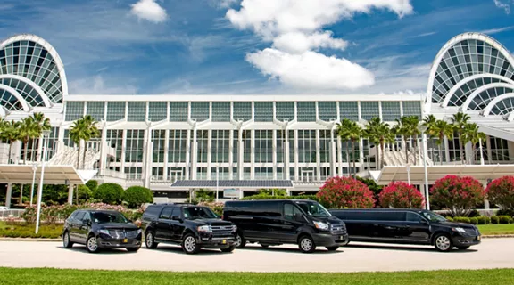 A fleet of 4 black vehicles, one small SVU, one large SUV, one passenger van and a limo, parked outside of a large building