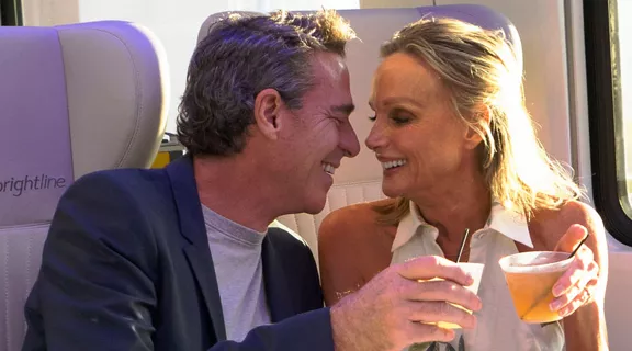 A man and woman toast their drinks while riding a Brightline train.