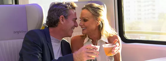 A man and woman toast their drinks while riding a Brightline train.
