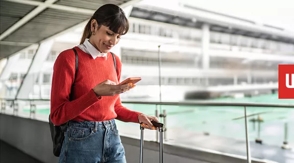 A woman on the go with her rolling suitcase in one hand and looking down at her phone in the other, a WallyPark Airport Parking logo over-layed to the right.