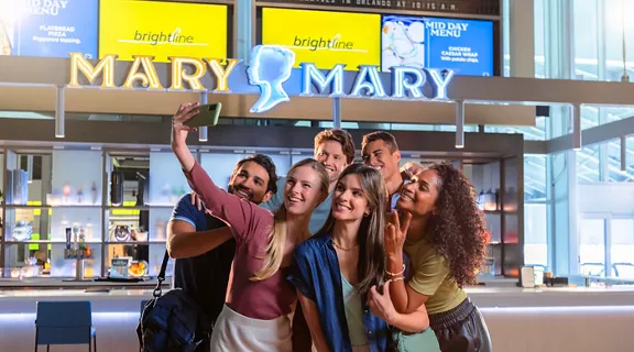 A group of people taking a selfie in front of Brightline Orlando Station's Mary Mary bar