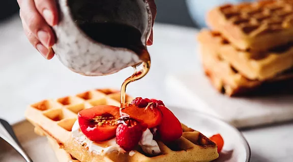 Close-up of a female hand pouring maple syrup on waffle. Woman serving sweet and tasty breakfast.