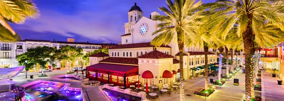 A vibrant Mediterranean-style plaza is captured at dusk, featuring a central building with a large clock tower and red awnings. Tall palm trees frame the scene, while illuminated fountains and pools add colorful light in the foreground. Surrounding structures with arches and balconies suggest a lively shopping or dining district. The sky glows in shades of blue and purple, enhancing the festive, resort-like atmosphere.