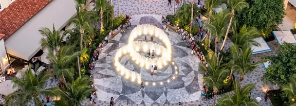 A circular plaza with a bright illuminated fountain is seen from above, surrounded by palm trees and people. The patterned stone pavement radiates outward from the central water feature, creating a decorative geometric design. Visitors sit and walk around the plaza, giving the scene a lively, social atmosphere. Warm lighting and lush greenery suggest a tropical resort or outdoor entertainment area at dusk.