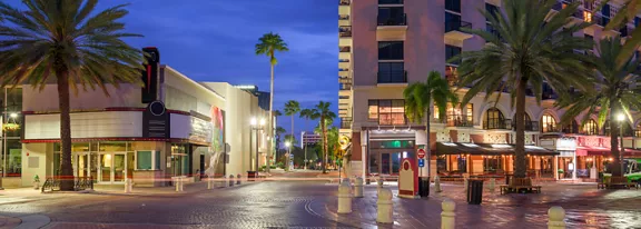 A quiet urban street scene is captured at twilight, lined with tall palm trees and mixed-use buildings. Restaurants and storefronts glow with warm lights, contrasting with the deep blue evening sky. Outdoor seating and pedestrian walkways suggest a lively yet relaxed downtown atmosphere. The scene feels calm and inviting, with no visible crowds.