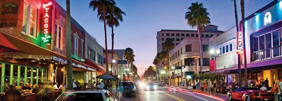 A lively downtown street scene is captured at dusk with cars driving along a central road lined by palm trees. Restaurants and shops on both sides of the street are illuminated by bright neon signs, including a vertical sign reading TACOS on the left. Pedestrians sit at outdoor tables and walk along the sidewalks, adding to the bustling urban atmosphere. The sky transitions from blue to purple, enhancing the vibrant, colorful nightlife mood.