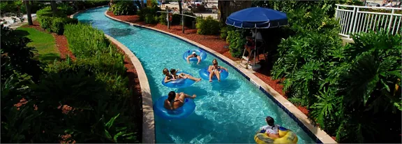 Several people float on inflatable tubes along a winding lazy river pool. The waterway is surrounded
