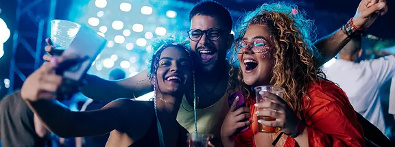 A small group of friends are at a music festival taking a selfie together with excited expressions on their faces.