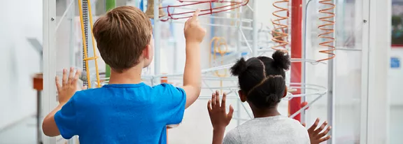Two young children stand closely together, viewing an interactive mechanical exhibit behind glass. The boy points toward the colorful tubes and tracks while the girl looks on with curiosity. Bright coils and pathways suggest a playful science or museum environment. The scene feels educational and engaging, emphasizing discovery and hands-on learning.