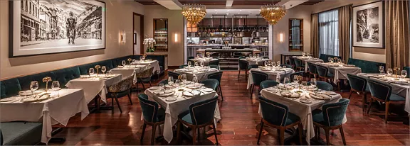 Upscale restaurant dining room with neatly set tables and upholstered chairs is shown in a warm, inv