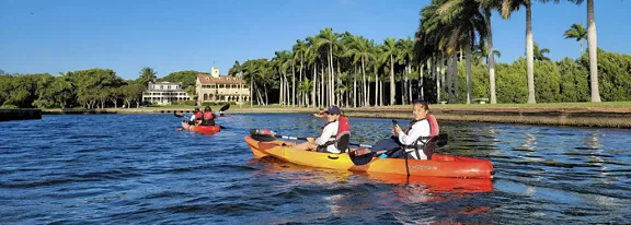Several people paddle colorful kayaks along a calm waterfront canal lined with tall palm trees. A large house or resort building is visible in the background under a clear blue sky. The kayakers appear relaxed and engaged in outdoor recreation on the water. The scene conveys a warm, tropical atmosphere ideal for leisure and sightseeing.