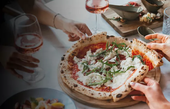 A freshly baked Neapolitan-style pizza topped with arugula and soft cheese sits on a wooden board at the center of a table. 