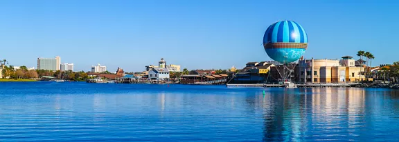 A large blue striped hot air balloon rises above a waterfront entertainment district. Modern low-rise buildings line the shore, with palm trees and distant high-rise structures visible against a clear blue sky. Calm water in the foreground reflects the balloon and surrounding architecture, creating a bright and colorful scene. The overall atmosphere is sunny, relaxed, and inviting.