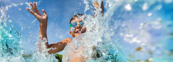 A child is captured mid-splash in clear blue water, with arms raised playfully. The close-up perspec