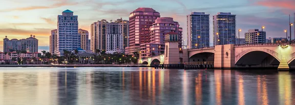 A vibrant city skyline rises along a calm waterfront at dusk. Tall modern buildings line the shore, reflecting softly in the still water. An elegant bridge extends from the right side of the frame, connecting the city across the waterway. The sky glows with pastel hues of pink, orange, and blue, adding warmth to the urban scene.