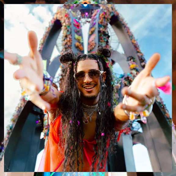 A young adult with long curly hair and sunglasses reaches toward the camera, smiling brightly. 