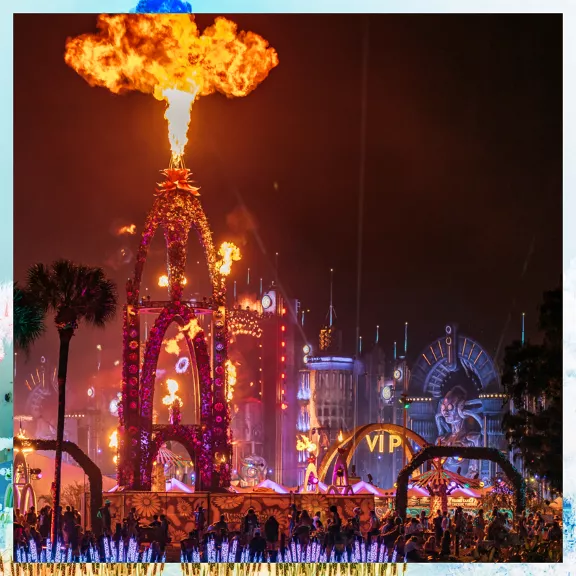 A vibrant nighttime festival scene features a towering structure shooting a large burst of fire into the sky. 