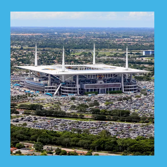 A large modern sports stadium is seen from above, surrounded by expansive parking lots filled with cars. 