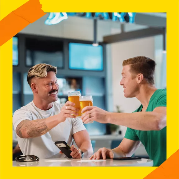 Two men sit at a modern bar counter, smiling as they clink glasses of beer.