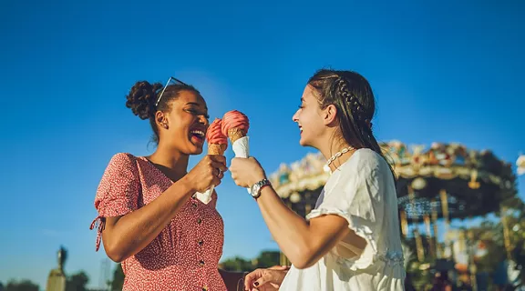 Two women standing in front of a carousel at a theme park on a sunny day, eating ice cream cones