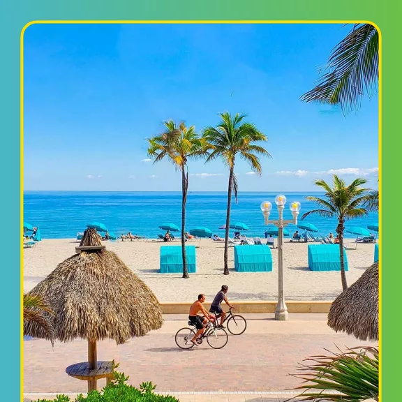 Two people ride bicycles along a paved beachfront promenade beside a sandy tropical beach.