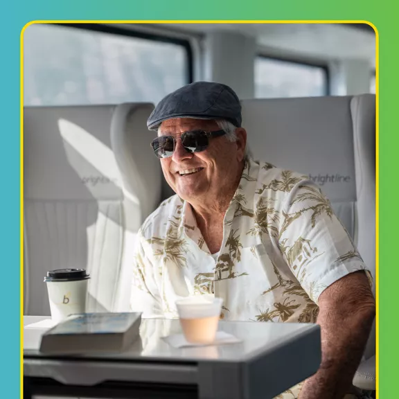 An older man wearing a patterned shirt, flat cap, and sunglasses sits comfortably in a modern train seat.