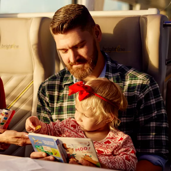 A bearded man in a plaid shirt reads a children’s book with a young child wearing a red bow headband. 