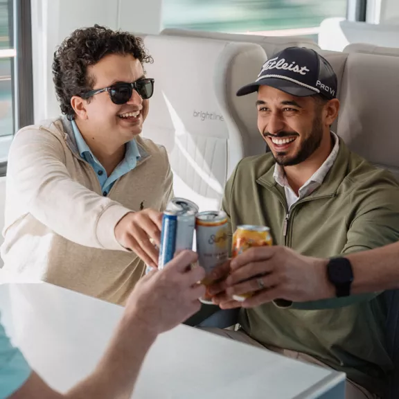 Two adult men sit together on a Brightline train, smiling as they clink aluminum cans with another person across the table. 