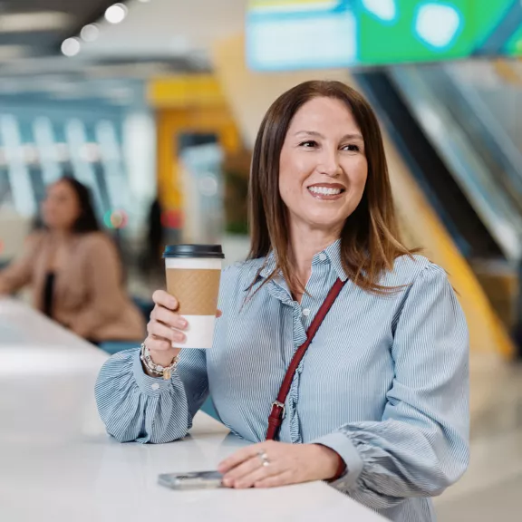 A woman sits at a bright counter holding a takeaway coffee cup and smiling.
