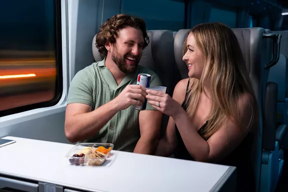 A young couple sits together on a modern train, clinking a can and a plastic cup in a casual toast. They share a small meal tray with snacks on the table between them. 
