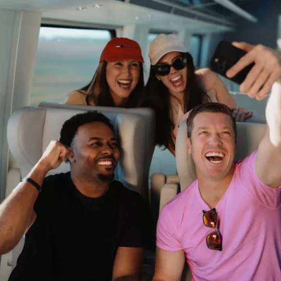 A group of four friends pose for a selfie while traveling on a modern train. Two people sit in the foreground, one wearing a bright pink shirt and sunglasses, the other in a dark shirt. Two more friends lean in from behind the seats, one wearing a red cap.