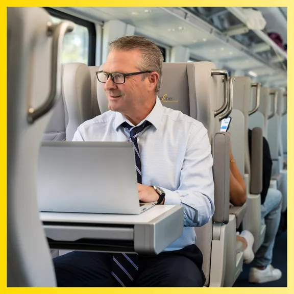 A middle-aged man in business attire works on a laptop while seated on a modern train. He wears a light blue dress shirt, striped tie, and glasses, and uses a fold-out tray table as his workspace. The train interior features gray reclining seats and bright natural light from the windows.
