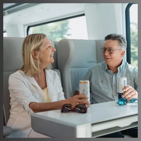 A mature couple sits together at a table on a modern train, smiling and talking while holding canned beverages. 