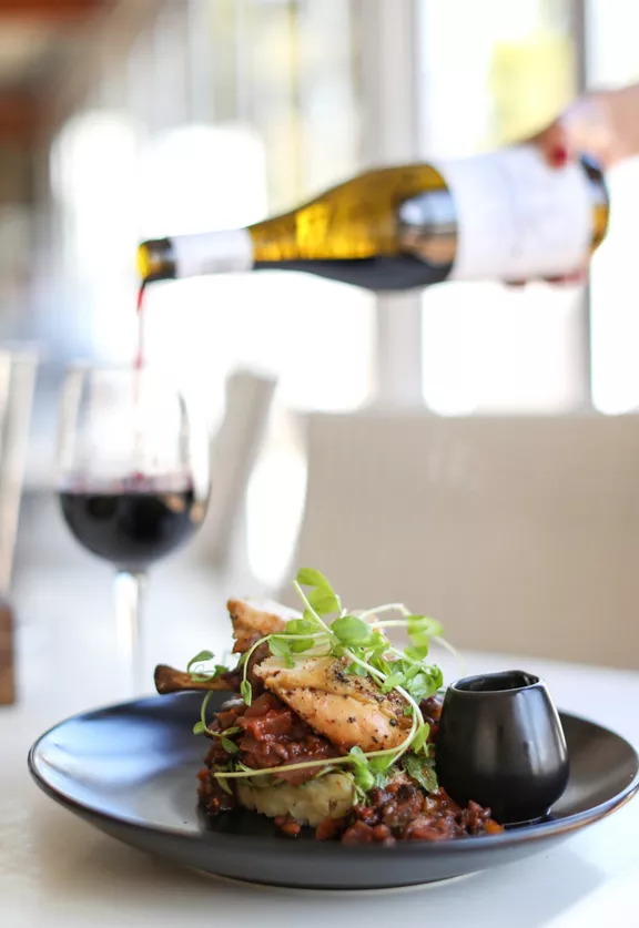 A dinner plate of seafood topped with greens with a glass of red wine being poured in the background
