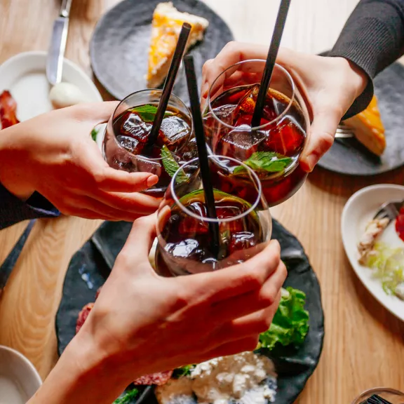 Three people raise round glasses filled with iced dark beverages and straws in a celebratory toast.