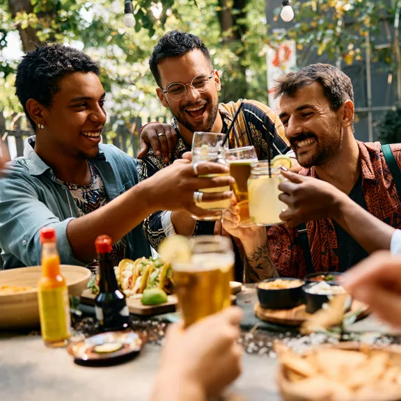 A group of friends raise their glasses in a cheerful toast at an outdoor table. The setting appears to be a casual restaurant or backyard gathering with trees and sunlight in the background. 