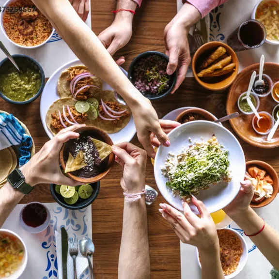 A group of people gather around a wooden table, sharing a variety of Mexican dishes. Tacos, rice, sauces, and lime wedges are arranged in bowls and plates as hands reach in to serve and pass food.