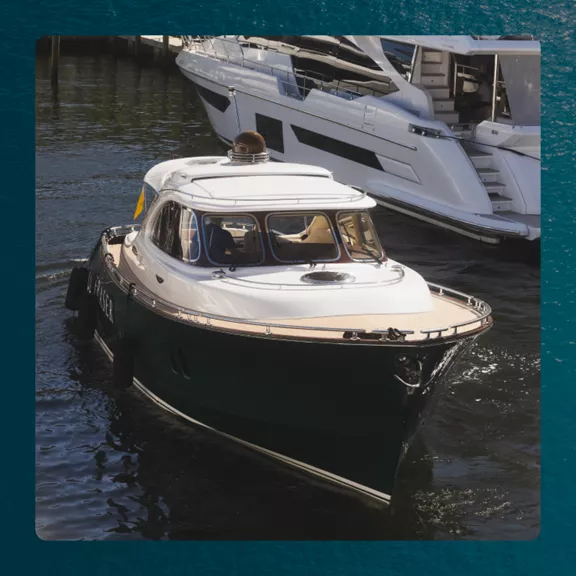 A compact motor yacht with a dark hull and light deck cruises slowly through a marina. 