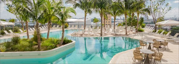 A large, curving swimming pool is surrounded by tall palm trees and lush greenery at a tropical resort. Numerous white lounge chairs and umbrellas line the pool deck, suggesting a relaxing vacation setting. In the foreground, small round tables and chairs provide seating near the water. The background shows city buildings and a bright blue sky with scattered clouds, adding an urban coastal atmosphere.