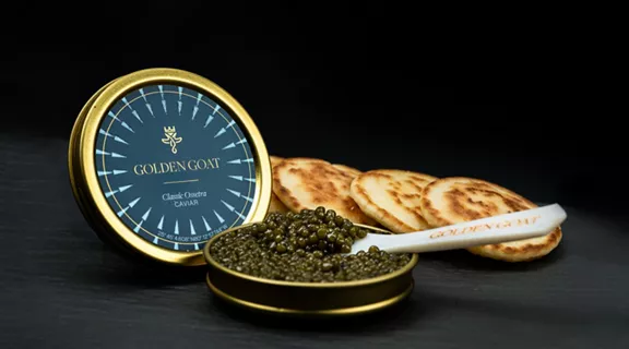 An opened tin of Golden Goat caviar with a serving spoon and round crackers