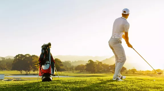 A man swinging a golf club on a course at sunrise.