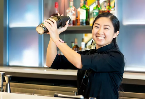 A smiling bar tender mixing a drink in mid air behind a bar