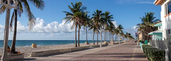A wide coastal boardwalk runs parallel to a sandy beach lined with tall palm trees. The calm ocean stretches to the horizon under a bright blue sky with scattered white clouds. Low-rise buildings and houses border the walkway on the right side, suggesting a relaxed seaside town. The scene feels peaceful and inviting, with clear weather and soft natural light highlighting the shoreline.